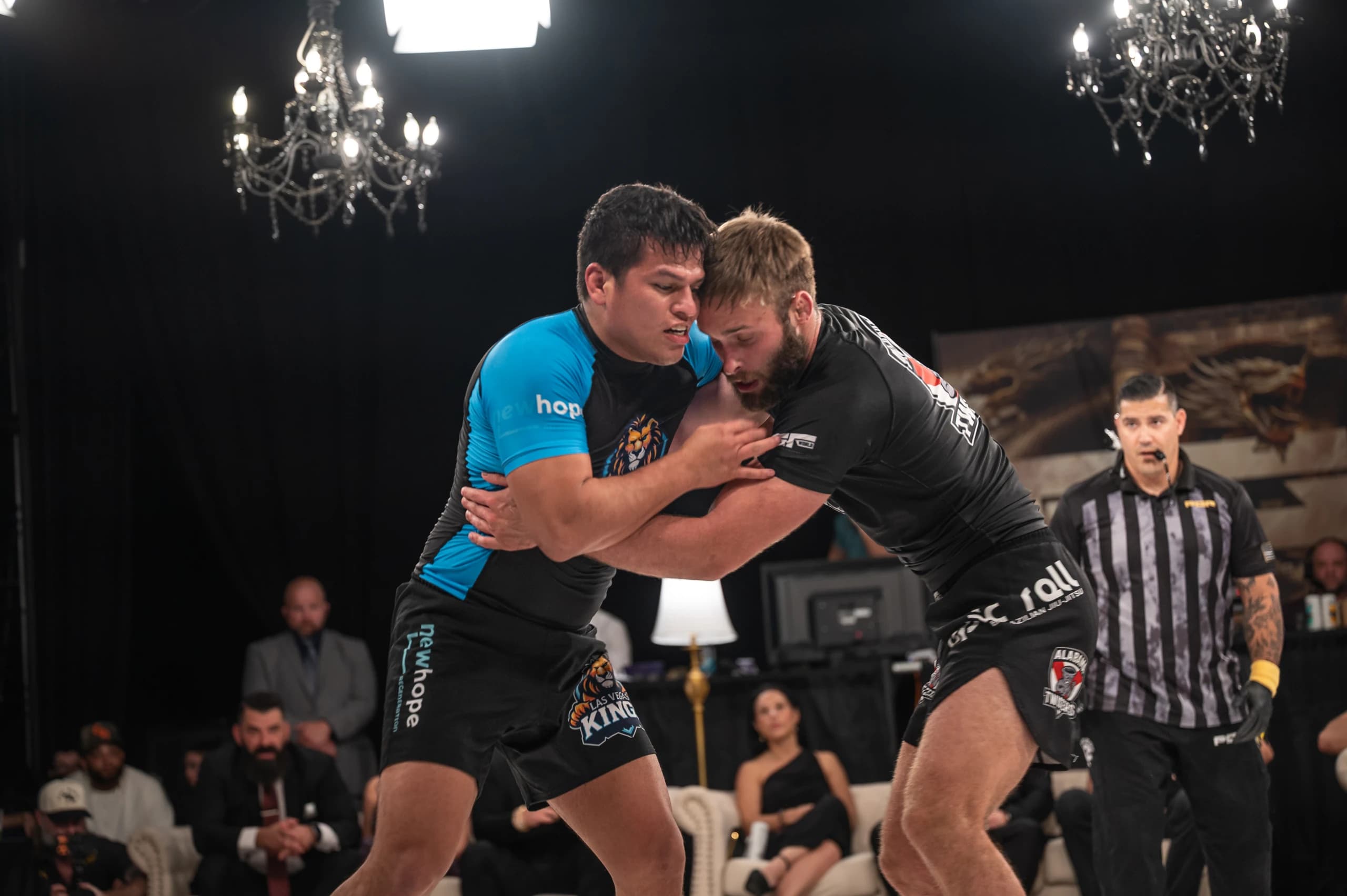 Las Vegas Kings and Alabama Twisters athletes grappling beneath chandeliers, with spectators seated at the edge of the PGF mat.